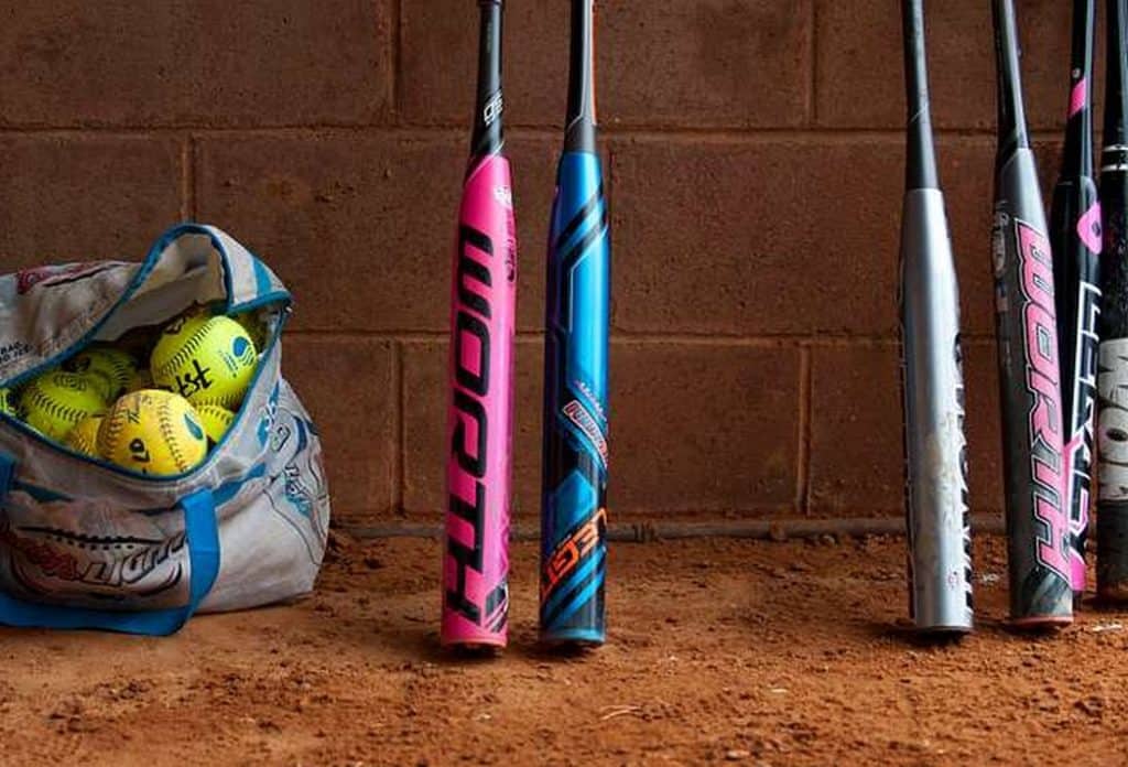 Bags of softballs and a couple of softball bats are lined up against a wall.